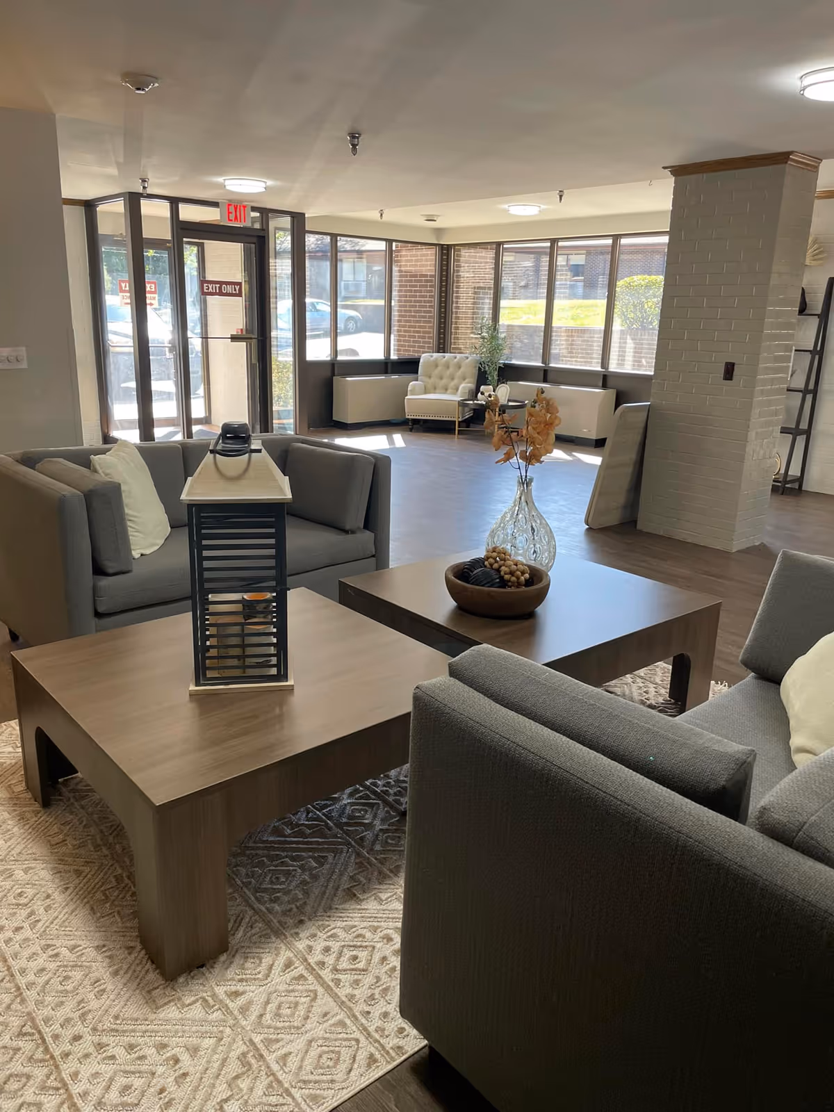 A bright and spacious senior living facility common area with large windows letting in natural light. The room features modern gray sofas with white pillows, two wooden coffee tables with decorative items including a lantern and a glass vase with dried flowers. In the background, there is a white armchair near the windows and a brick column in the middle of the room. The entrance door with an exit sign is visible on the left side.