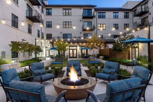 Outdoor courtyard area of a senior living facility at dusk with a central fire pit surrounded by six cushioned chairs. String lights are hung above, creating a warm ambiance. The courtyard is landscaped with small trees, shrubs, and patio umbrellas, and the multi-story building with balconies and large windows surrounds the space.