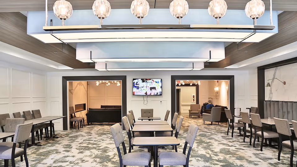 A spacious, well-lit dining area with multiple tables and chairs arranged neatly. The room features modern light fixtures hanging from the ceiling, patterned carpet flooring, and two open doorways leading to adjacent lounge areas with sofas and armchairs. A flat-screen TV is mounted on the wall between the doorways.