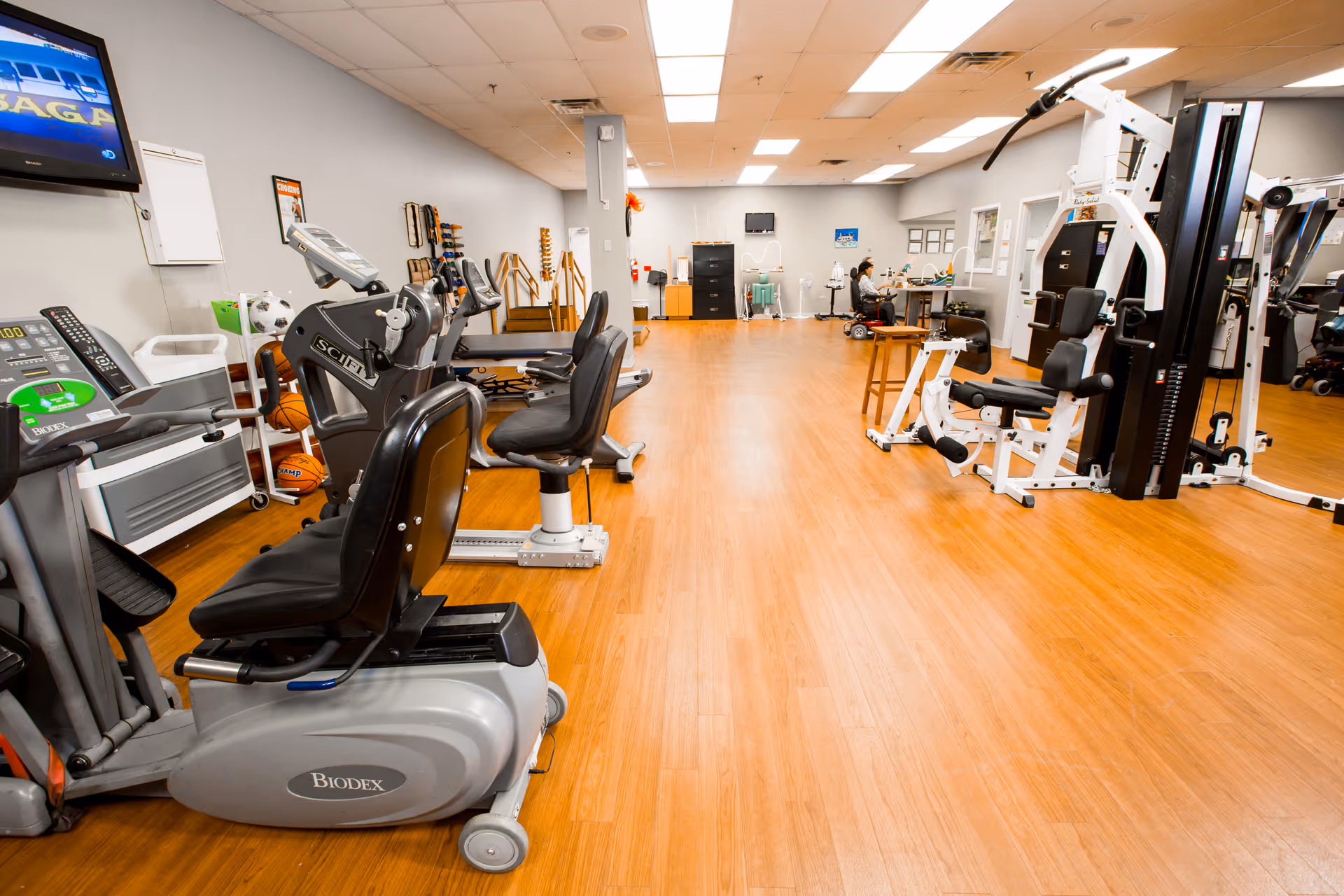 A spacious rehabilitation gym room with various exercise equipment including stationary bikes, weight machines, and therapy tools. The room has wooden flooring, bright overhead lighting, and a television mounted on the wall. A person is seated at a desk in the background near medical equipment.