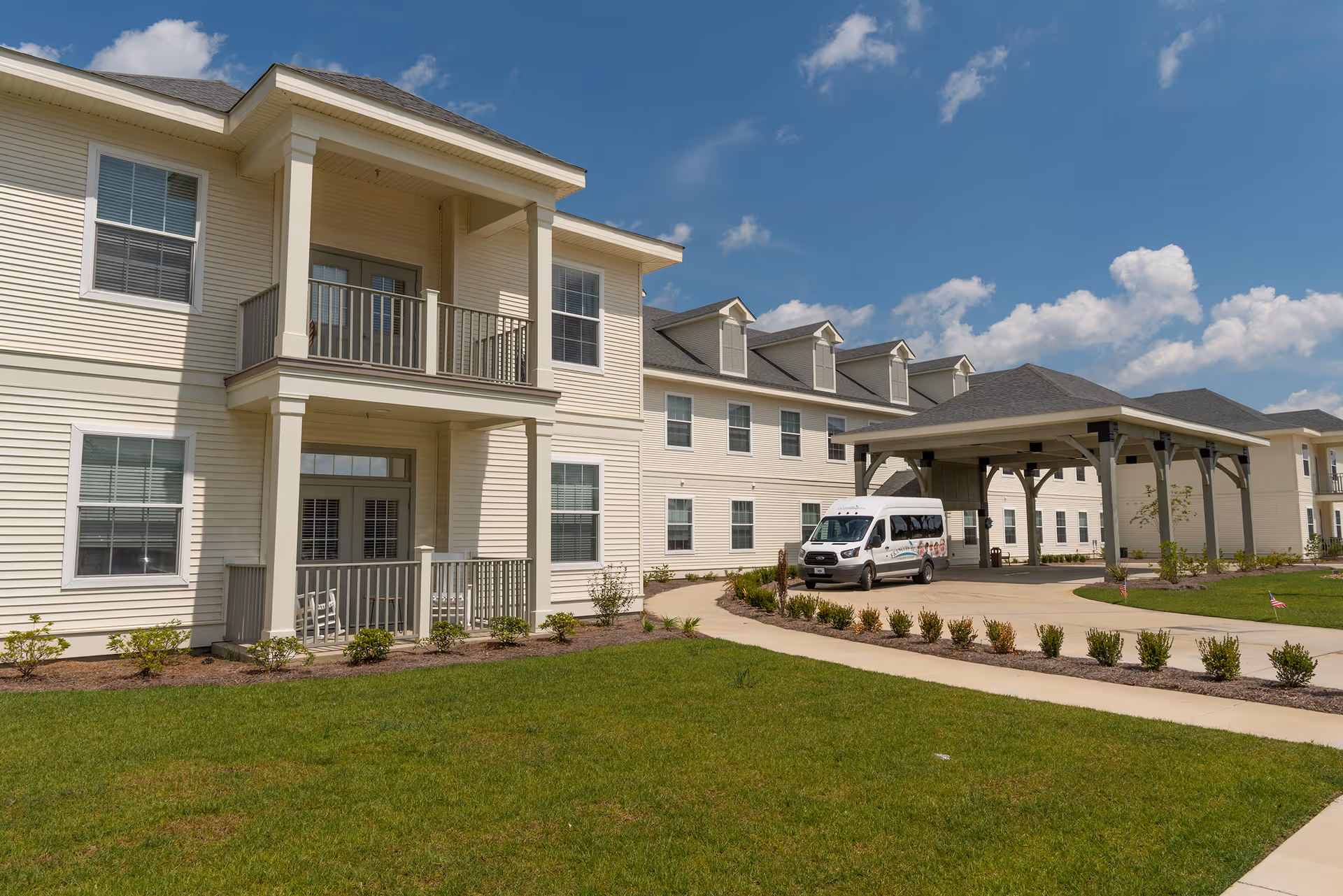 Exterior view of a senior living facility building with beige siding, multiple windows, and balconies. A covered entrance area is visible with a white shuttle van parked underneath. The foreground shows a well-maintained lawn and landscaped shrubs under a partly cloudy blue sky.