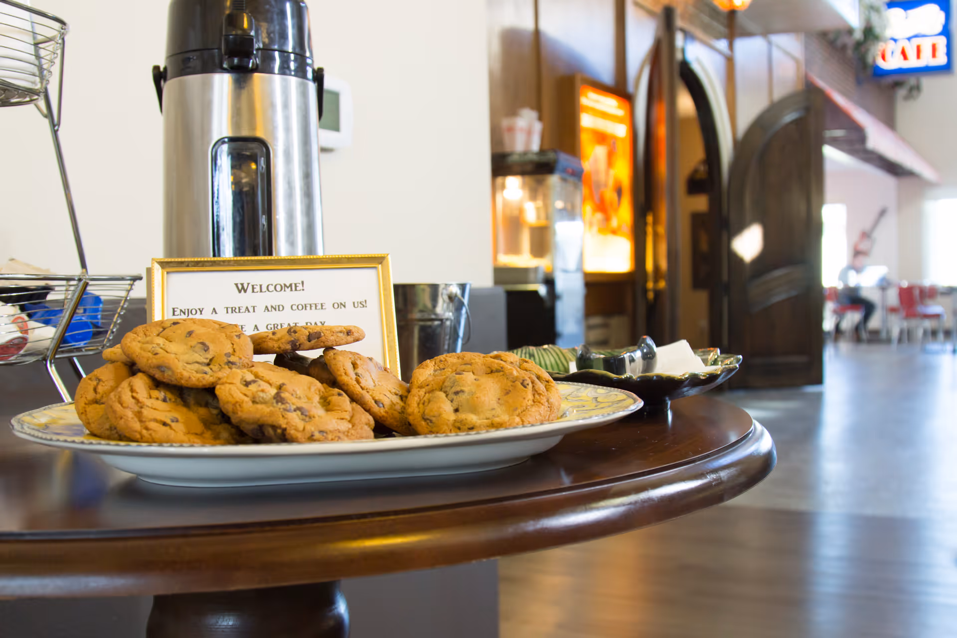 A plate of chocolate chip cookies on a wooden table with a coffee dispenser behind it. A small framed sign invites guests to enjoy a treat and coffee. The background shows an open doorway leading to a room with tables and chairs.