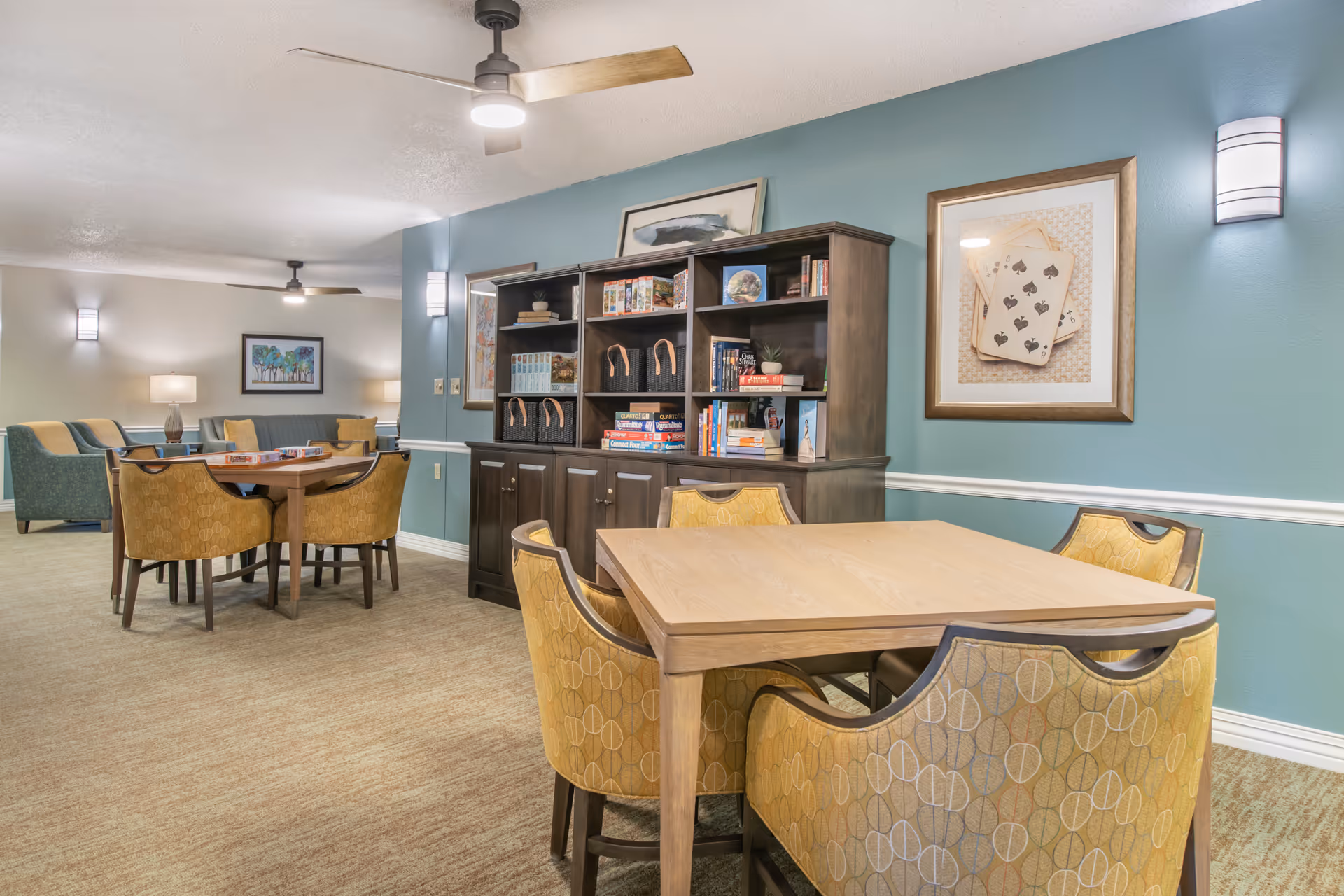 Well-lit common activity room with wooden tables and yellow upholstered chairs, bookshelves and framed artwork on teal walls.