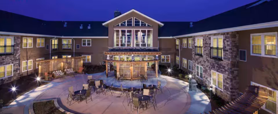 Nighttime courtyard of an assisted living facility with a central gazebo, outdoor seating, and a surrounding two-story building with lit windows.