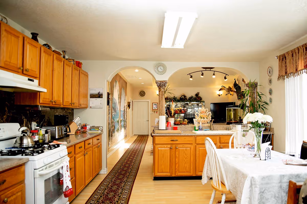 A bright and spacious kitchen and dining area with wooden cabinets, a white stove, and a countertop with various kitchen items. The dining table is covered with a white tablecloth and has a vase with white flowers. The room features a long patterned rug leading to a hallway with wall murals and a clock above the archway. The background shows a living area with plants, a TV, and decorative items.