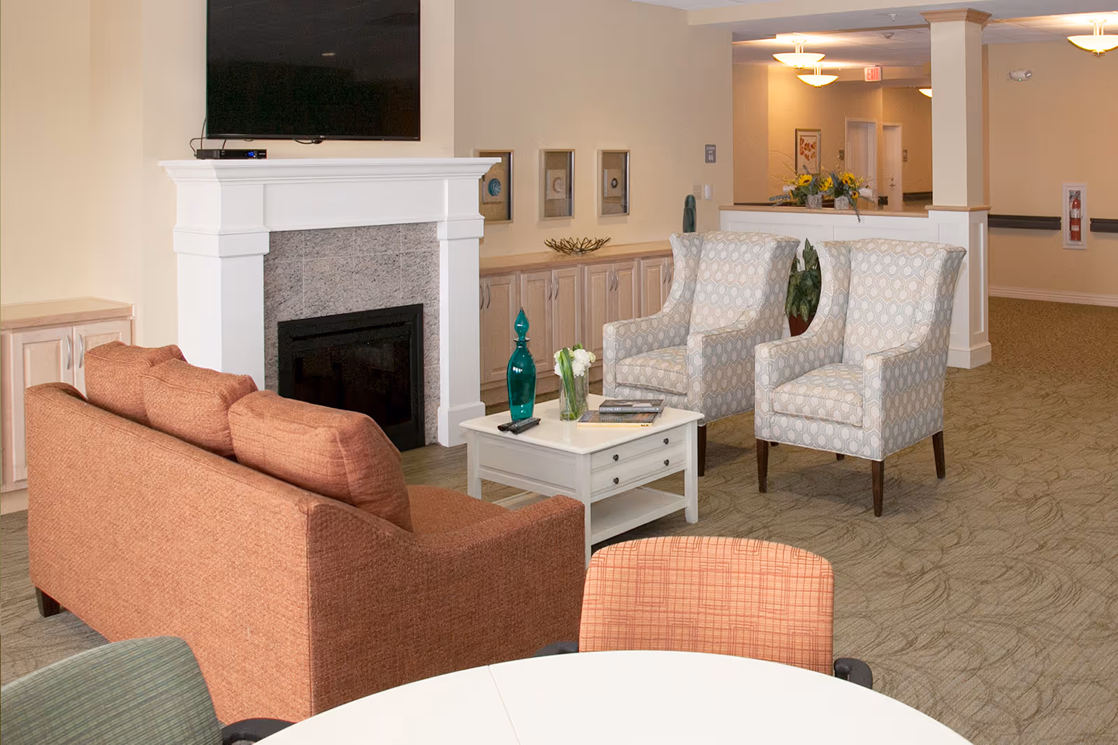A cozy living room area in a senior living facility featuring a burnt orange sofa, two patterned armchairs, a white coffee table with decorative items, a fireplace with a mounted flat-screen TV above it, and built-in cabinets along the wall. The room has soft lighting and a carpeted floor with a subtle leaf pattern.