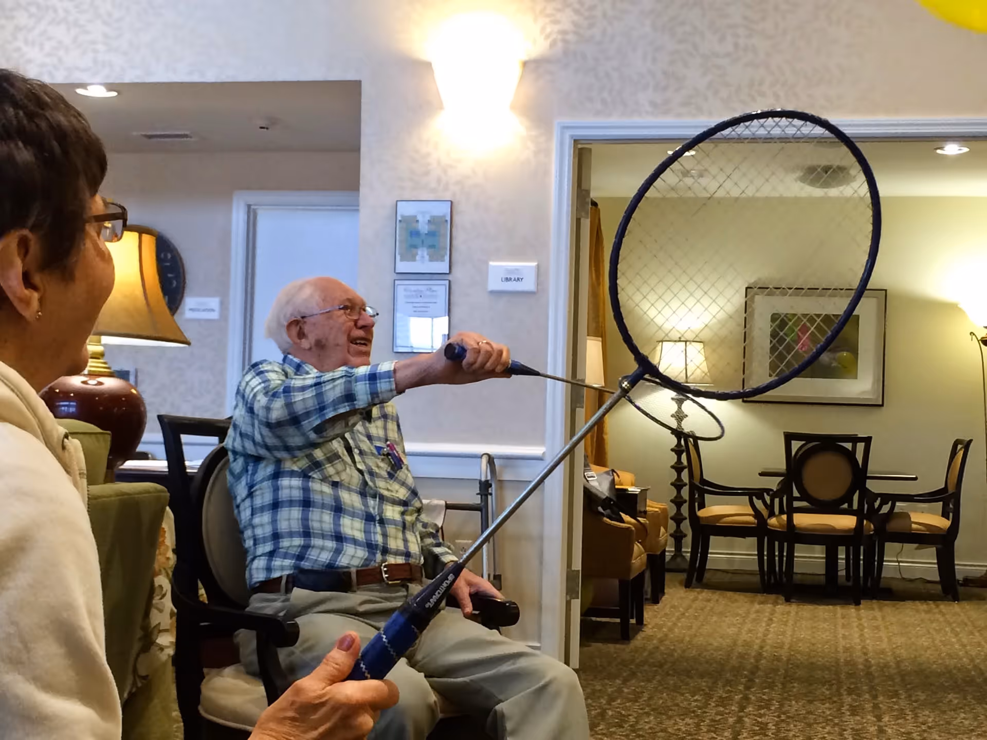 An elderly man sitting in a chair indoors, smiling and holding a badminton racket, with a woman sitting nearby watching him. The room has warm lighting, a lamp, and a dining area with chairs and a table in the background.