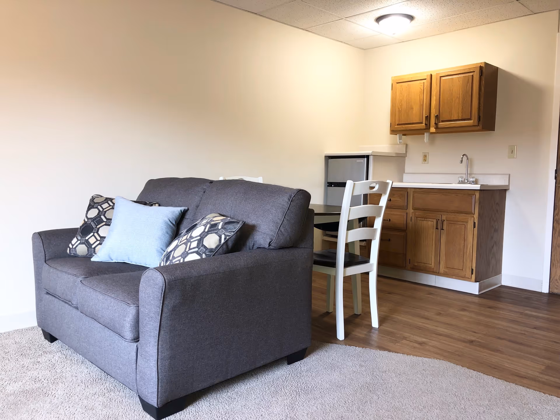 A small living area with a gray loveseat adorned with three decorative pillows, positioned on a carpeted floor. Adjacent to the loveseat is a wooden floor section with a small dining table and two white chairs. In the background, there is a kitchenette with wooden cabinets, a countertop, a sink, and a mini refrigerator. The walls are plain and light-colored, and a ceiling light fixture is visible.