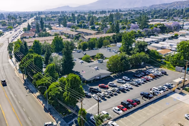 Aerial view of Claremont Care Center showing a large building complex surrounded by trees and greenery, with a parking lot filled with cars adjacent to the building. The facility is located near a main road with visible traffic and power lines, and residential areas and hills are visible in the background.