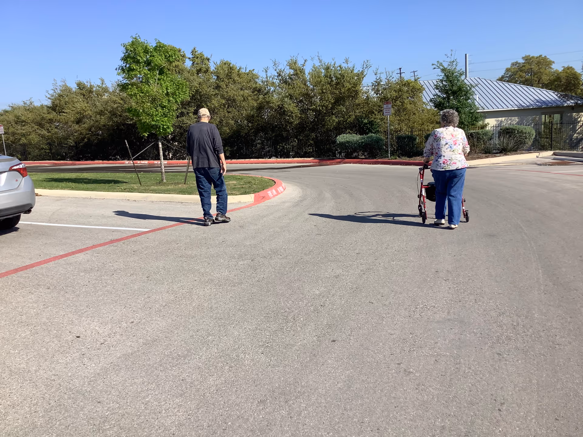 An elderly woman using a walker and an elderly man walking on a paved road near a parking area with trees and bushes in the background under a clear blue sky.
