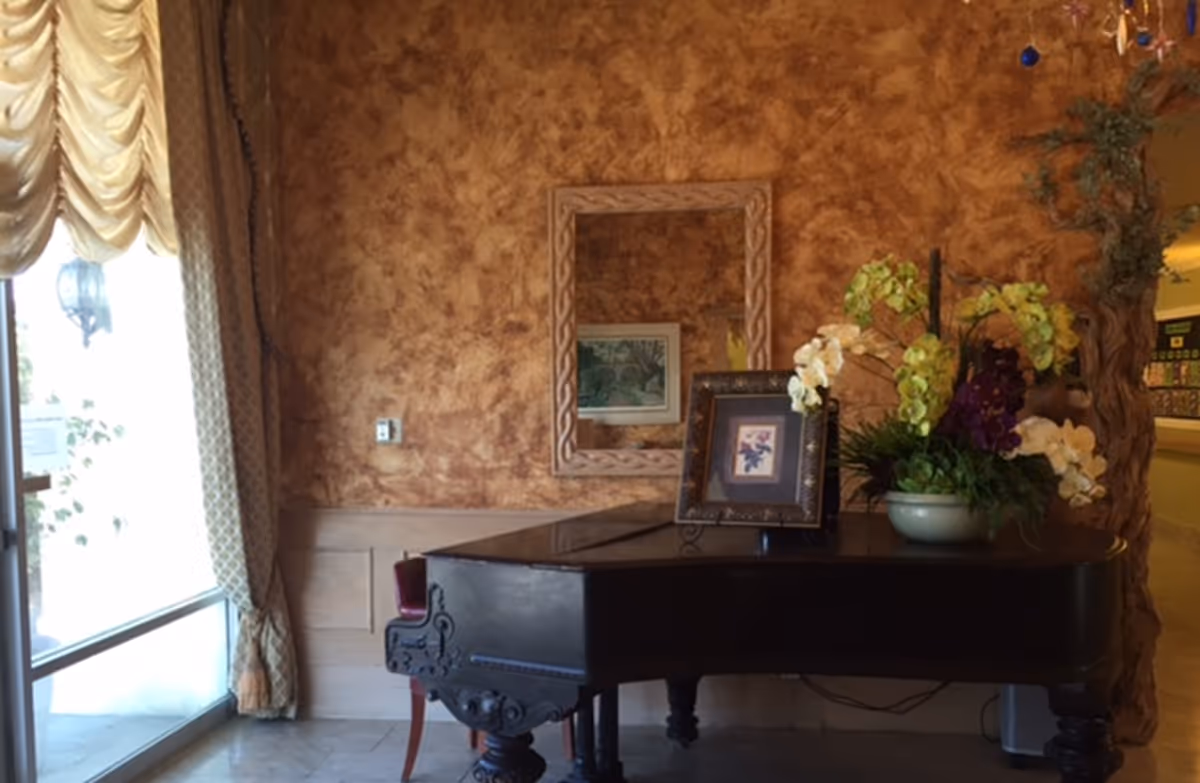 An elegant interior corner featuring a black grand piano adorned with a framed picture and a floral arrangement. The wall behind has a textured brown finish with a decorative mirror reflecting a framed artwork. To the left, there is a window with draped curtains allowing natural light to enter.