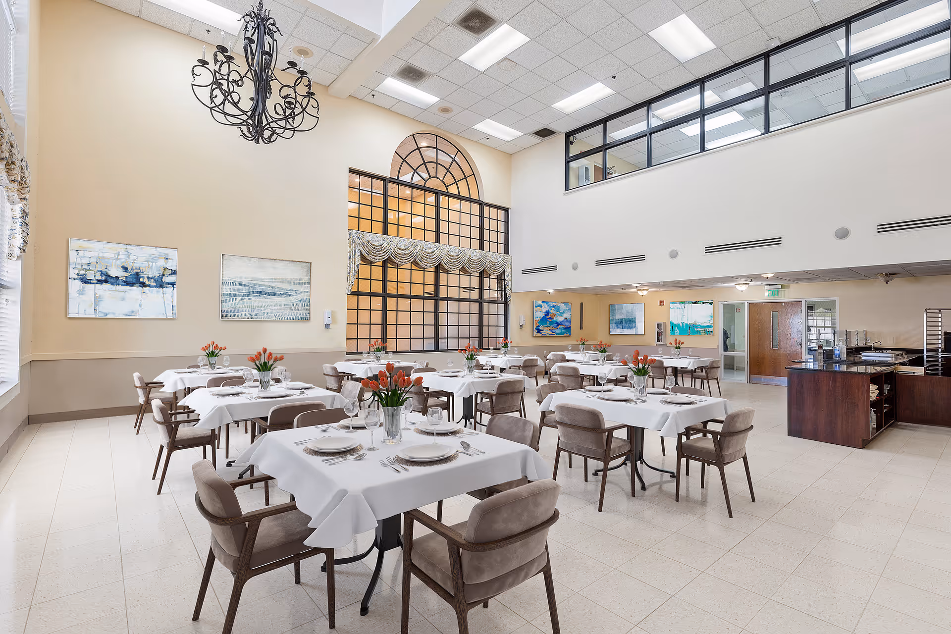Bright, high-ceiling dining room with neatly set tables and chairs, a chandelier, and large arched windows.