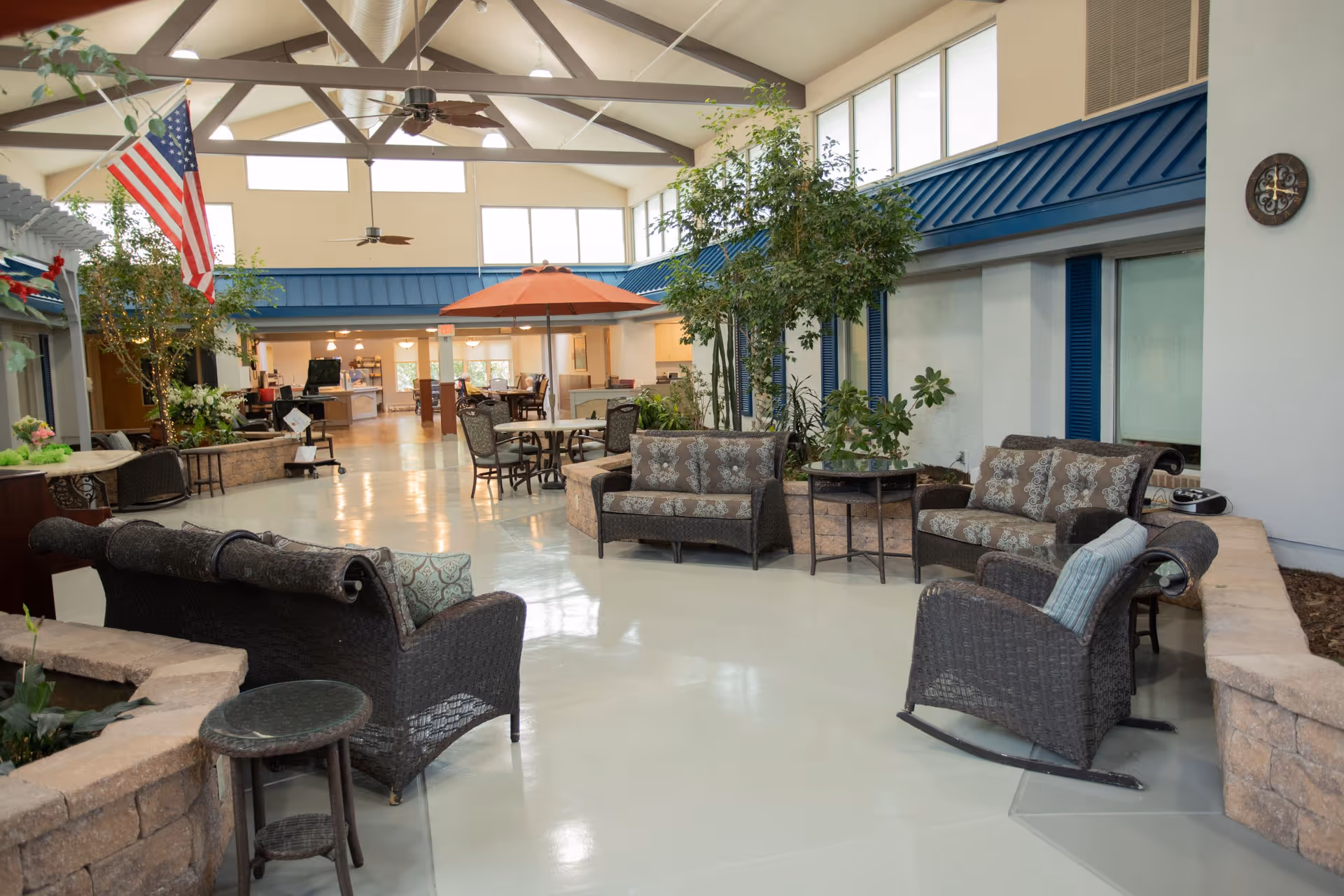 Indoor common area with wicker sofas and chairs arranged around small tables, potted plants, and an American flag hanging from the ceiling. The space has high ceilings with exposed beams and ceiling fans, large windows near the roof, and a dining area with tables and chairs in the background.