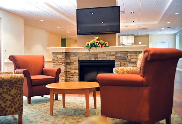 Comfortable seating area with red armchairs around a round wooden table facing a stone fireplace topped by a wall-mounted TV and a floral arrangement.