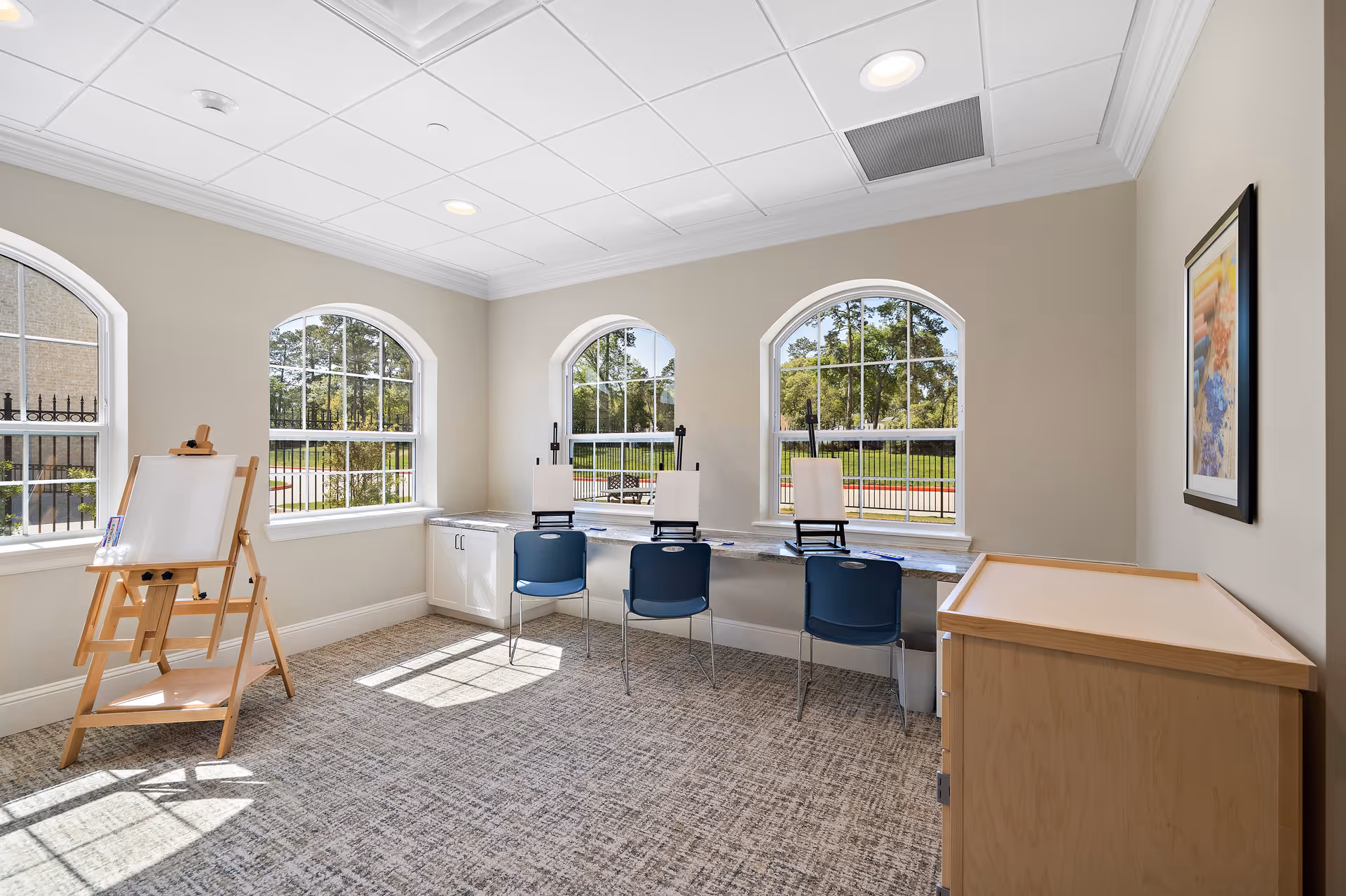 A bright art room with three large arched windows letting in natural light. There are three blue chairs in front of a long counter with three easels holding blank canvases. To the left, there is a wooden easel with a blank canvas. The room has beige walls, a patterned carpet, and a framed picture on the right wall.