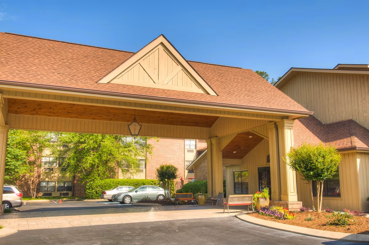 Covered porte-cochère entrance of a senior living community building with parked cars and landscaped beds.