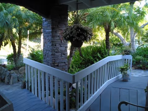 Covered outdoor porch with white railing, hanging basket, and lush tropical plants.