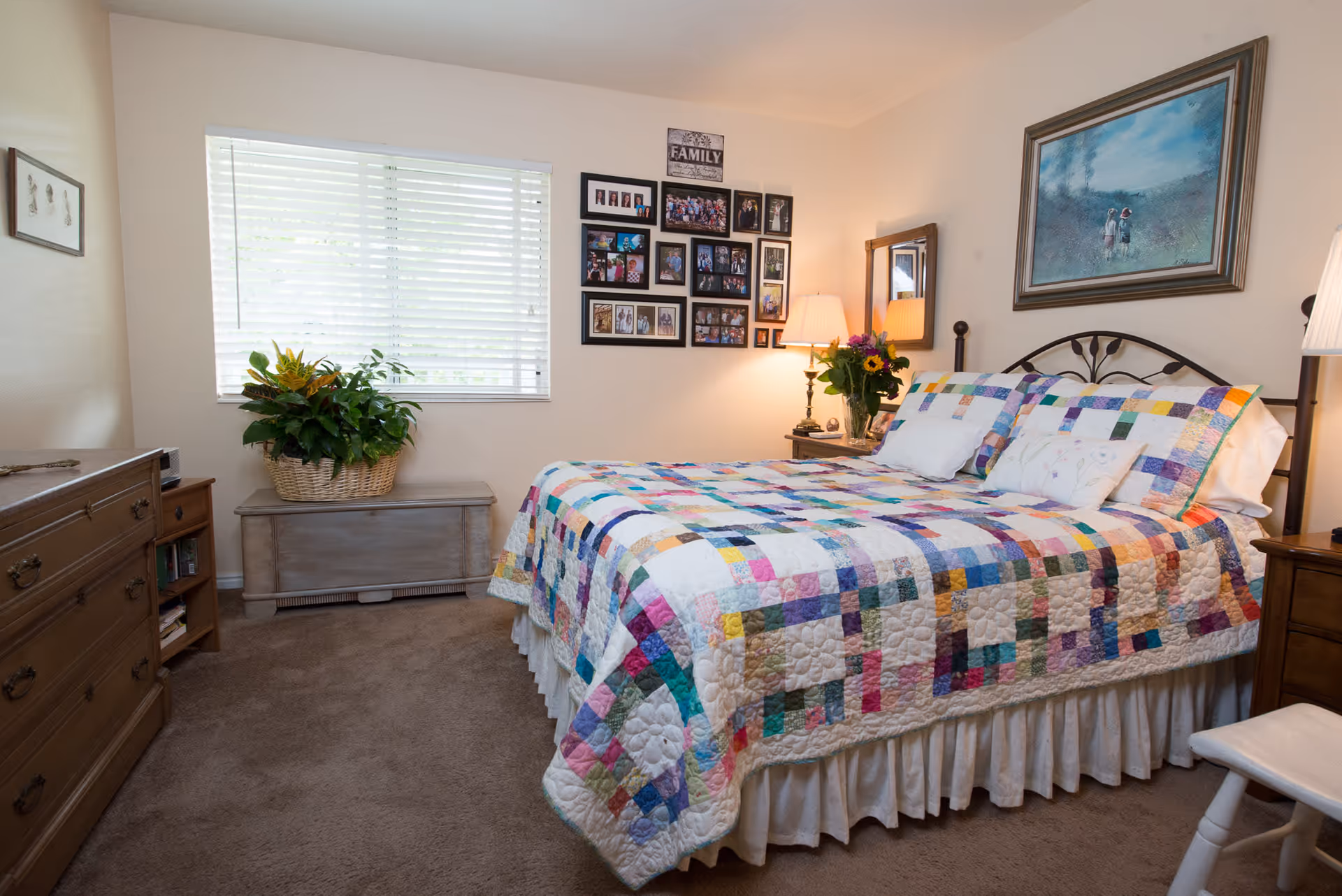 A cozy bedroom with a bed covered in a colorful patchwork quilt. The room has beige walls, a window with white blinds, a wooden dresser, a small chest with a basket of green plants on top, and a nightstand with a lamp and a vase of flowers. Family photos and artwork decorate the walls.