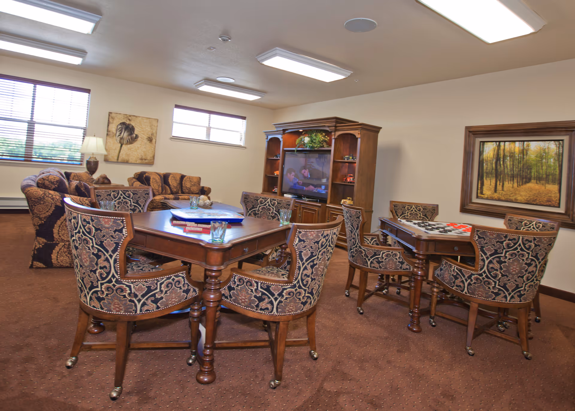 A cozy common area in a retirement living facility featuring two wooden game tables with patterned upholstered chairs. One table has a board game set up, and the other has a game in progress. In the background, there is a wooden entertainment center with a television and decorative items. A comfortable sofa and armchairs are arranged near two windows with blinds. The walls are decorated with framed artwork, and the room is carpeted with a warm brown color.
