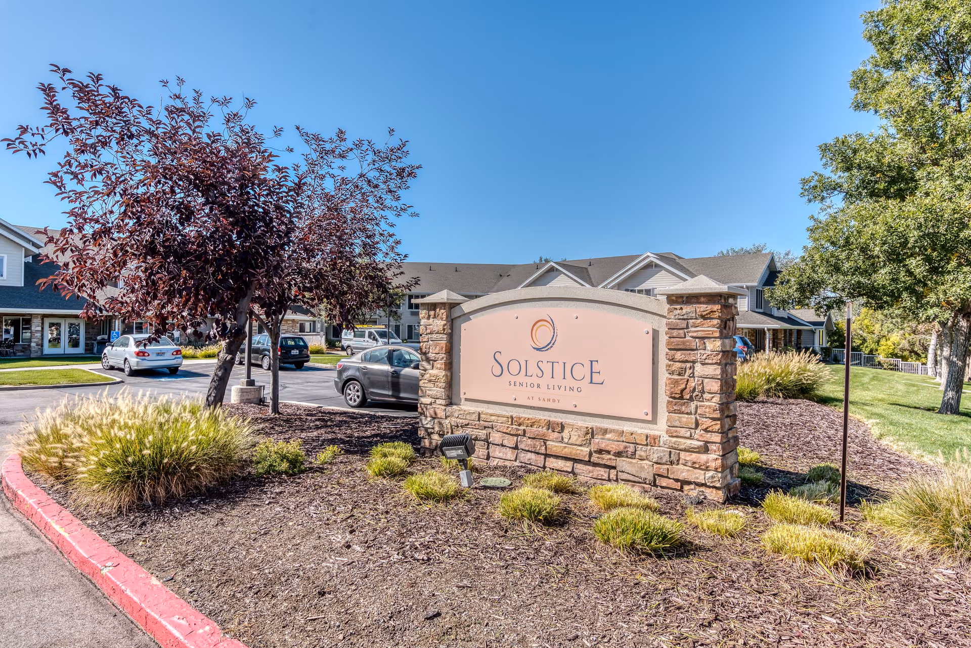 Outdoor view of the entrance sign for Solstice Senior Living at Sandy, surrounded by landscaped plants, trees, and a parking lot with cars. Residential buildings are visible in the background under a clear blue sky.