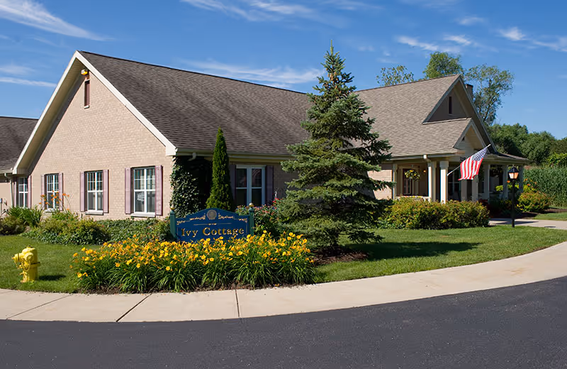Exterior view of a single-story brick building with a gabled roof, surrounded by green grass, yellow flowers, and trees. A blue sign in front reads 'Ivy Cottage.' An American flag is displayed near the entrance under a covered porch. The sky is clear and blue.
