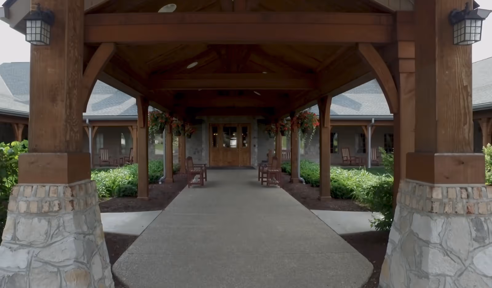 Covered wooden porte-cochere with stone pillars leading to the building's front doors, flanked by plants and rocking chairs.
