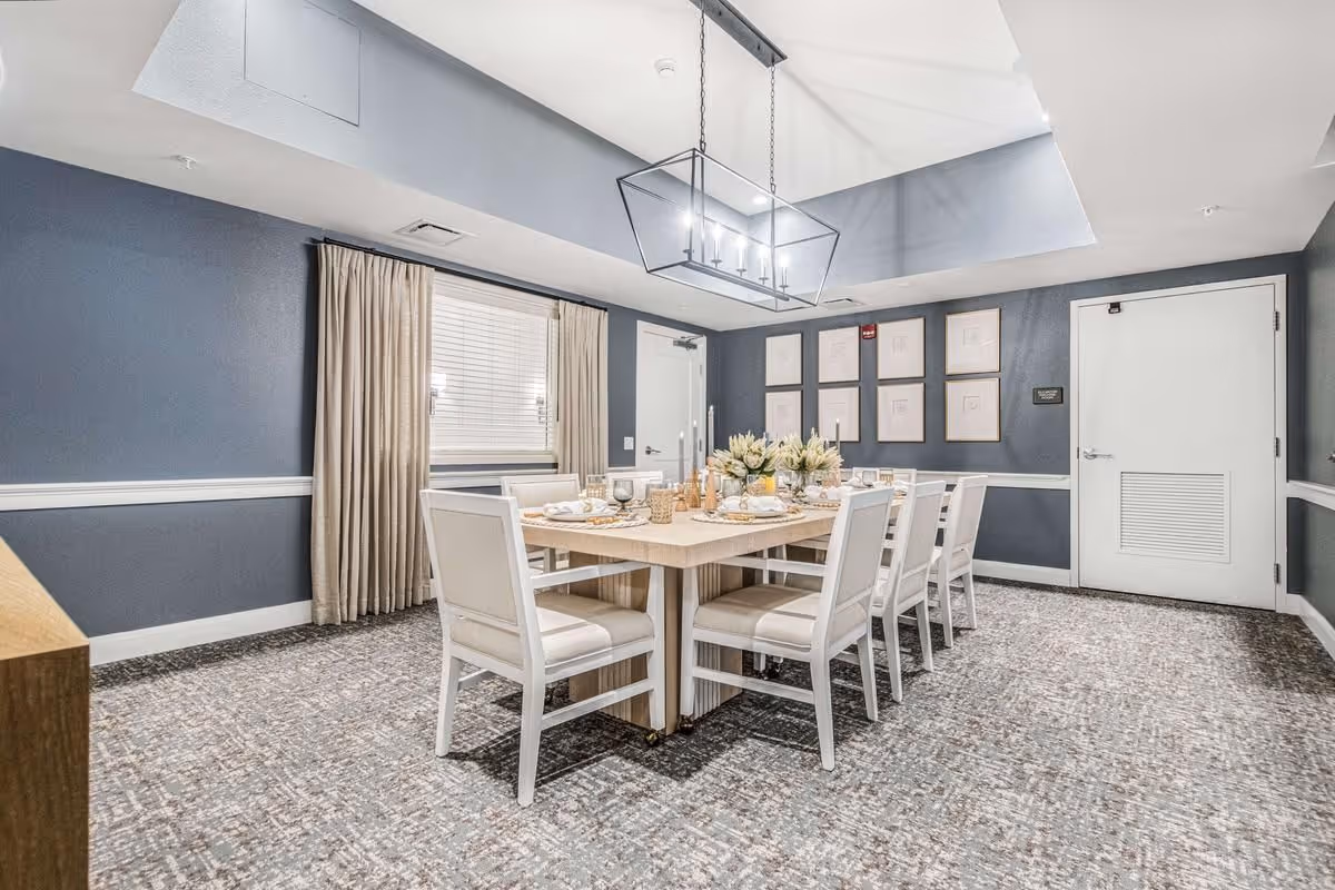 A formal dining room with a long table set for a meal, white chairs, and a rectangular chandelier under a recessed ceiling.