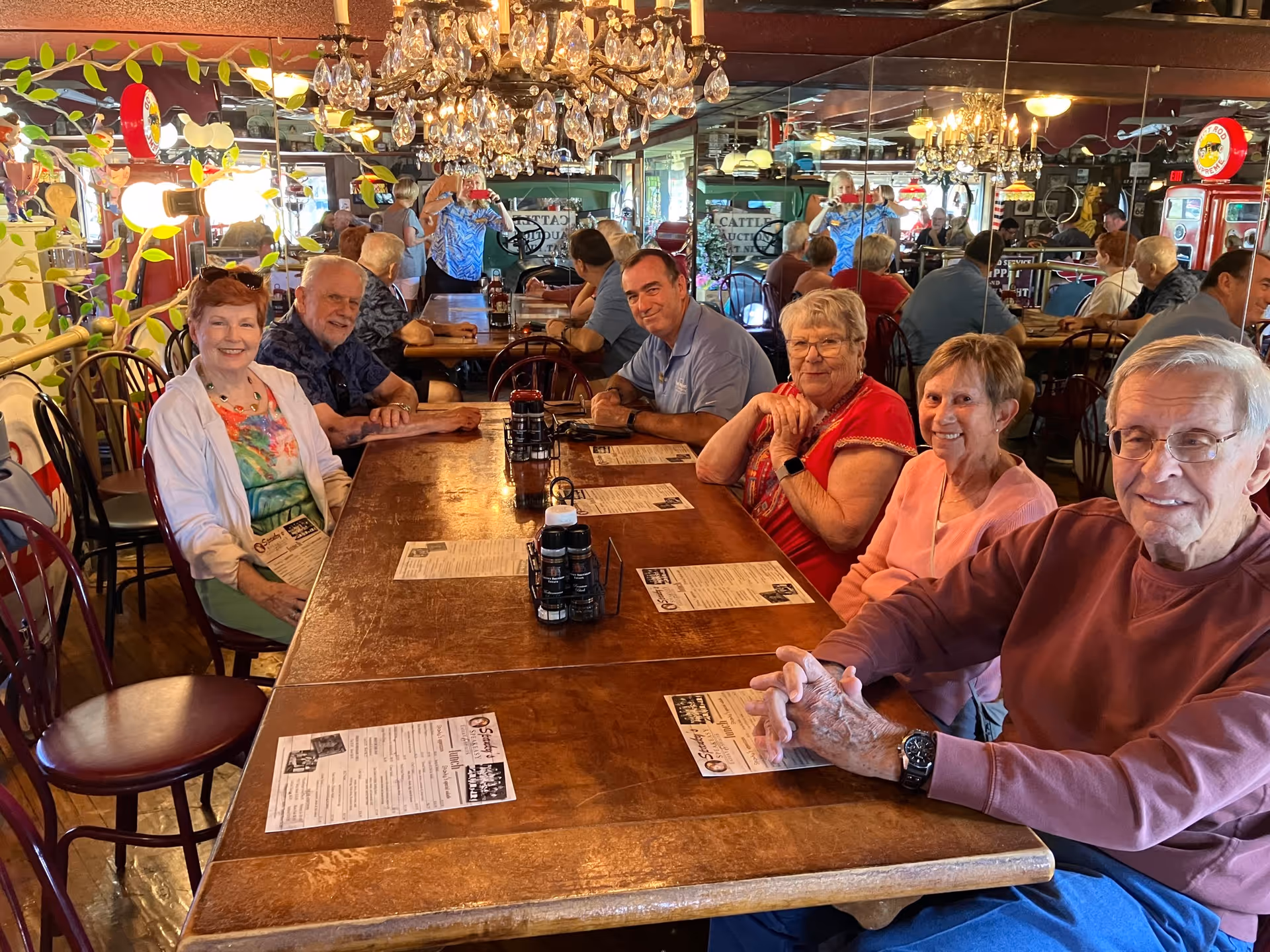 A group of six elderly people sitting around a wooden dining table in a restaurant, smiling and holding menus. The room has mirrored walls, chandeliers, and vintage decor including old signs and a red vending machine. Other diners are visible in the background.