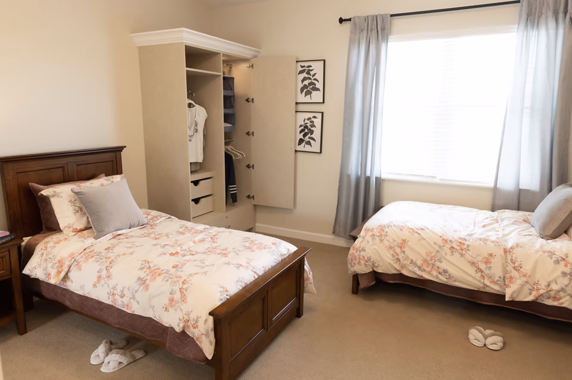 Bright bedroom with two twin beds in floral bedding, an open wardrobe, a bedside table, a large window with curtains, and slippers on the floor.