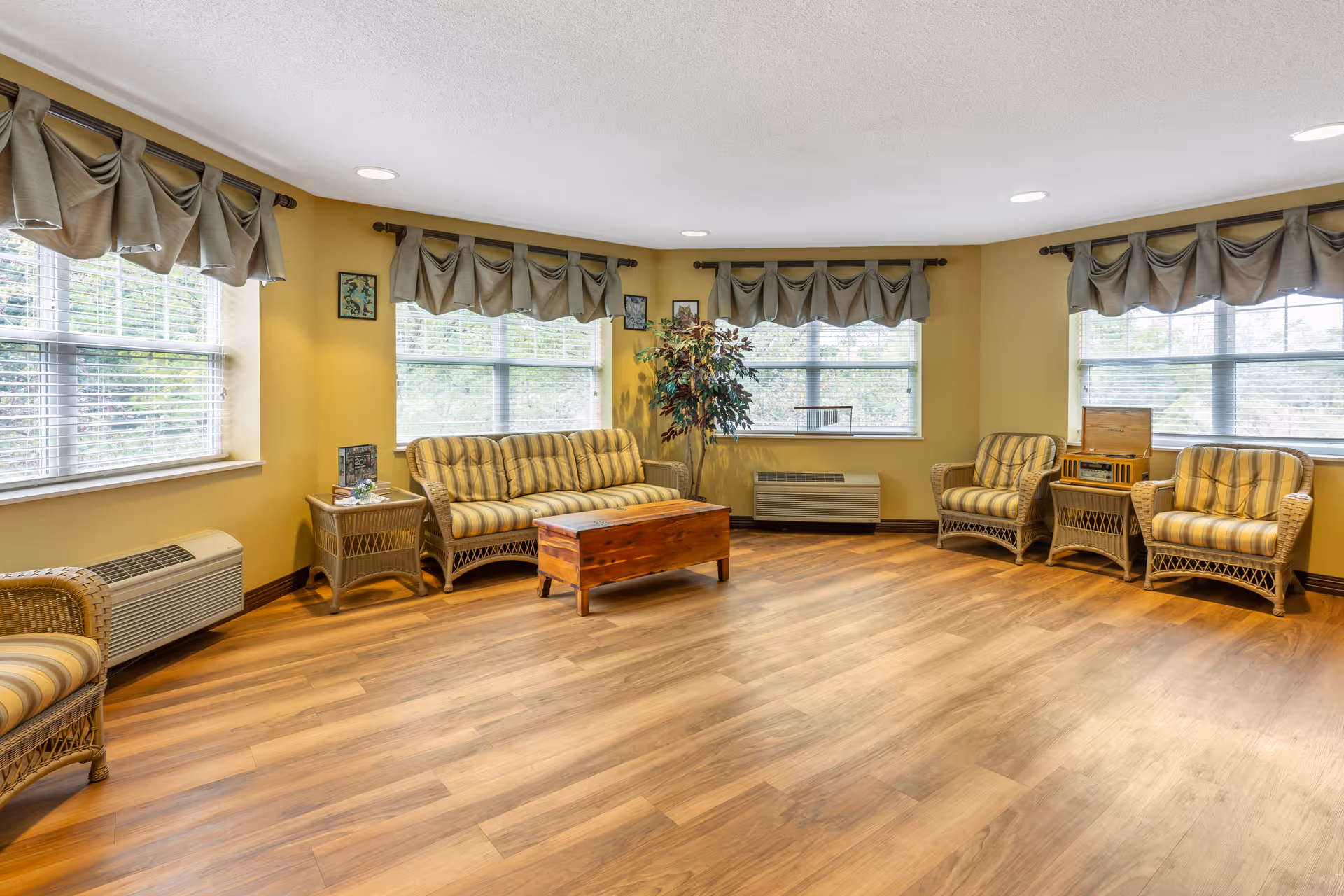 A bright and spacious living room with large windows covered by valance curtains. The room features wicker furniture including a striped sofa, three armchairs, a side table, and a wooden coffee table. There is a potted plant in the corner and a vintage-style radio on a wicker stand. The floor is wood, and the walls are painted a warm yellow.