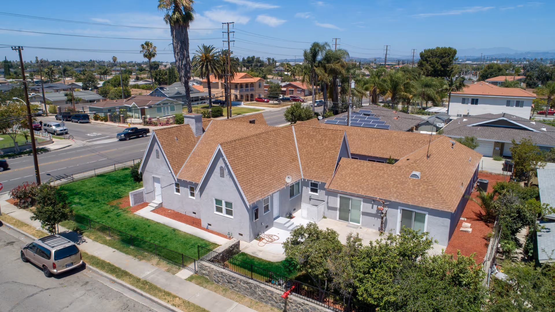 Aerial view of a single-story residential building with a brown shingle roof and light gray exterior walls, surrounded by a green lawn and trees. The building is located at a street corner in a suburban neighborhood with other houses, palm trees, and power lines visible in the background under a clear blue sky.
