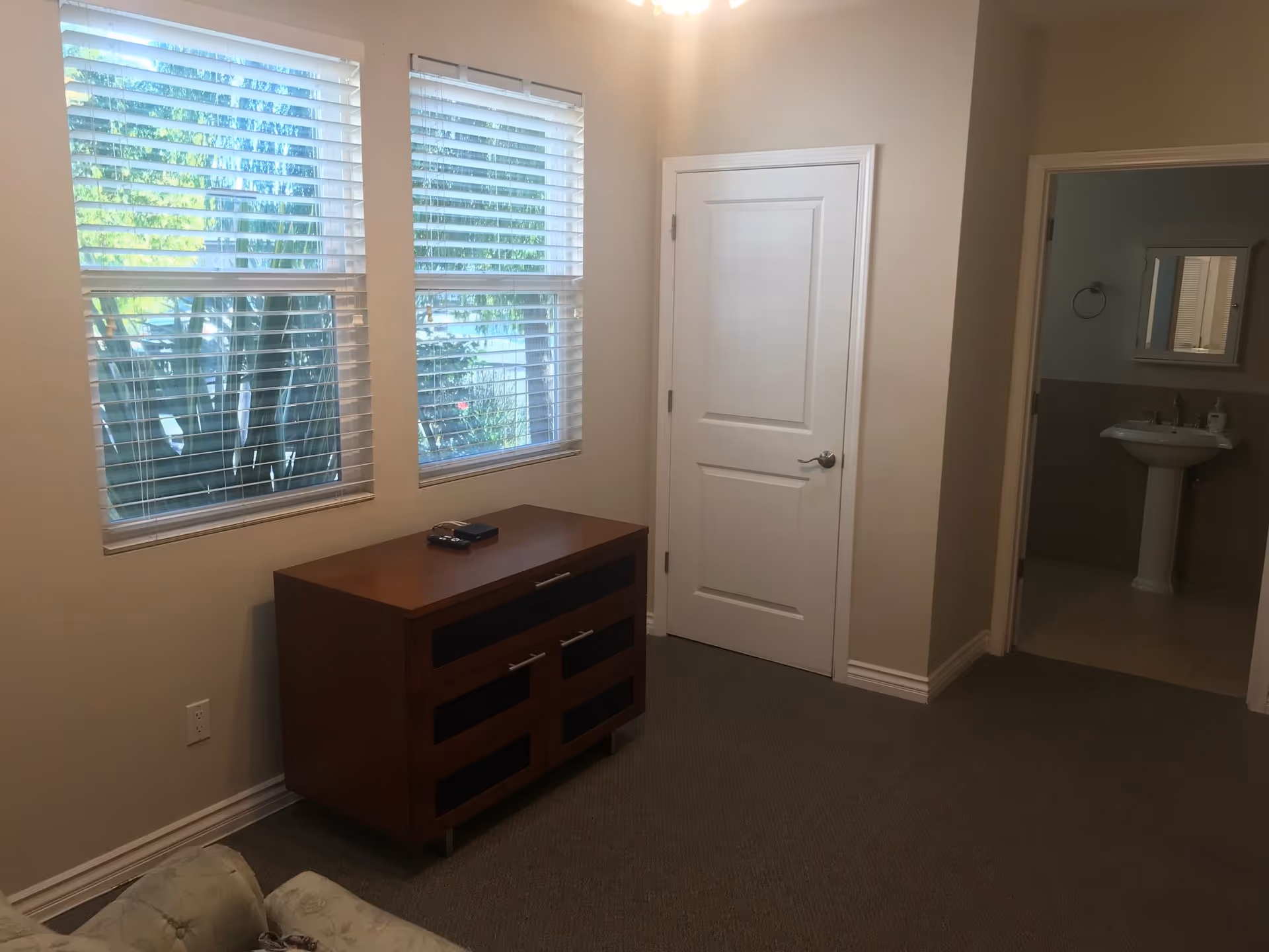 Small living room with two windows, a wooden dresser, an armchair corner, and a doorway leading to a bathroom.