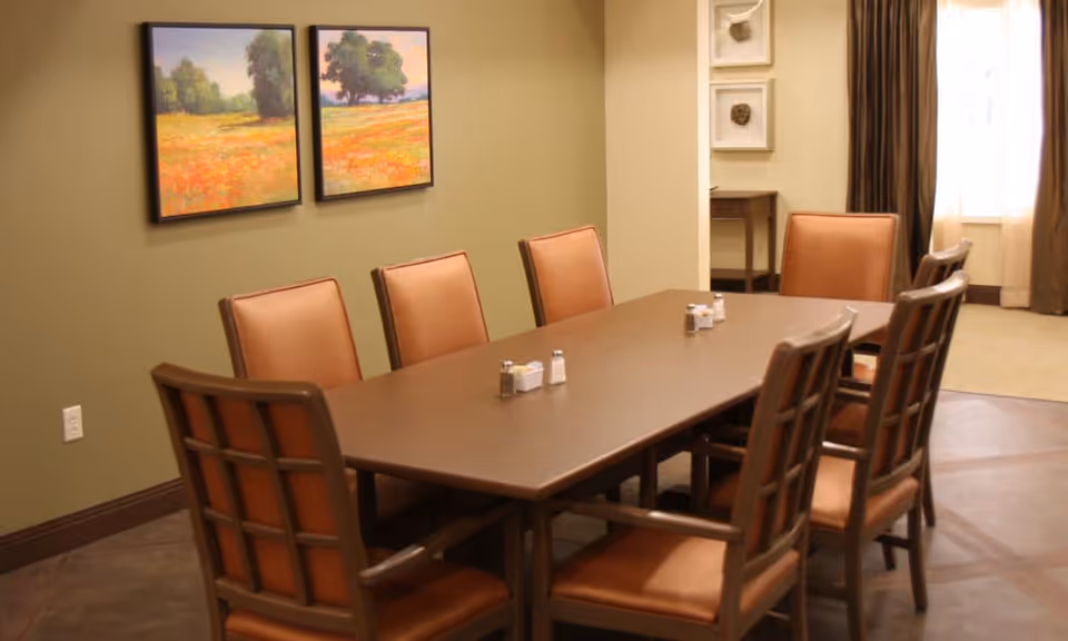 A dining room with a long rectangular wooden table surrounded by eight brown cushioned chairs. On the table are salt and pepper shakers. The walls are painted light green and decorated with two landscape paintings. In the background, there is a window with brown curtains and a small wooden table with framed artwork above it.