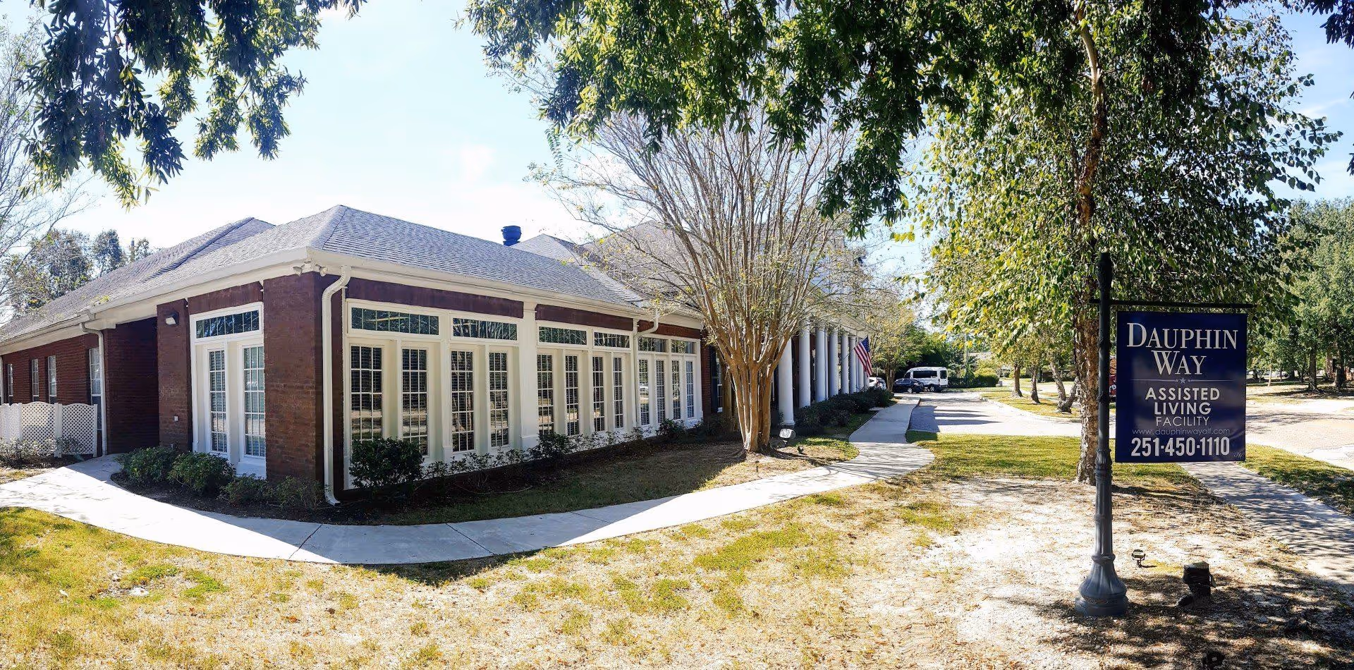 Exterior view of Dauphin Way Senior Living, an assisted living facility with a brick building featuring large windows and white trim. There is a sidewalk leading to the entrance, trees providing shade, and a sign displaying the facility's name, website, and phone number.