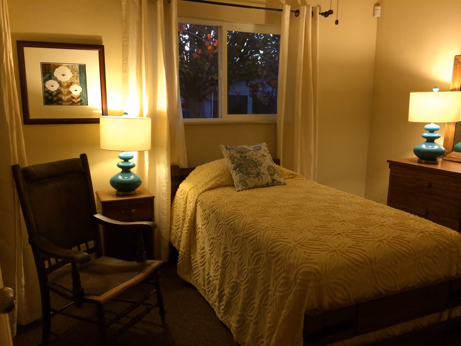 A cozy bedroom with a single bed covered in a textured yellow bedspread and a decorative pillow. There are two turquoise lamps on wooden nightstands on either side of the bed. A wooden chair is positioned next to one nightstand. A framed floral artwork hangs on the wall, and a window with cream-colored curtains shows a tree outside at dusk.