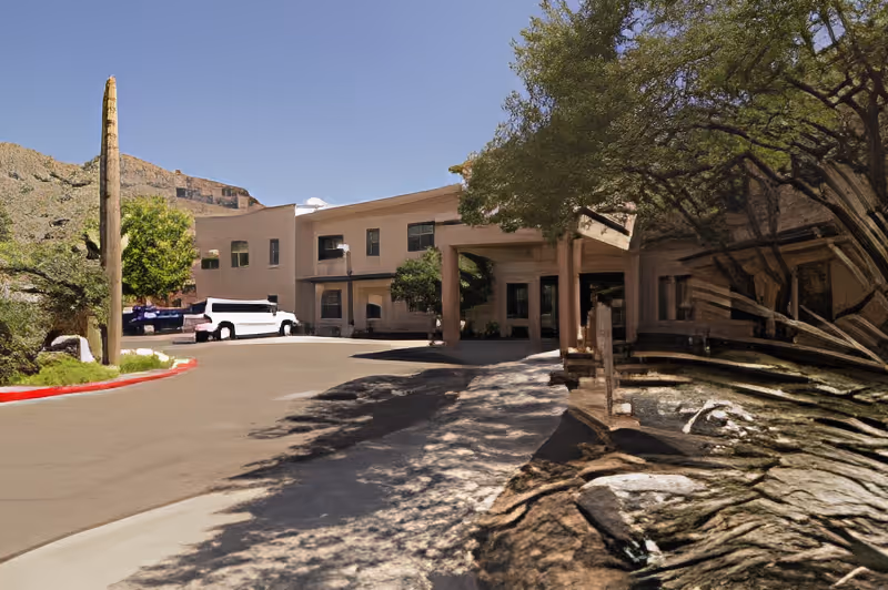 Exterior view of a senior living facility building with a covered entrance, a driveway, a white shuttle bus parked near the entrance, desert landscaping including a tall cactus and trees, and mountains in the background under a clear blue sky.