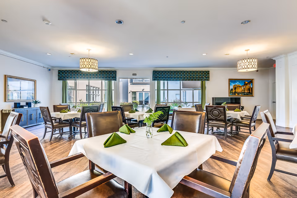 A bright and spacious dining room with several tables covered in white tablecloths and set with green folded napkins and small vases with green plants. The room has large windows with green curtains and valances, wooden floors, and modern light fixtures hanging from the ceiling. There are framed paintings on the walls and comfortable wooden chairs around the tables.