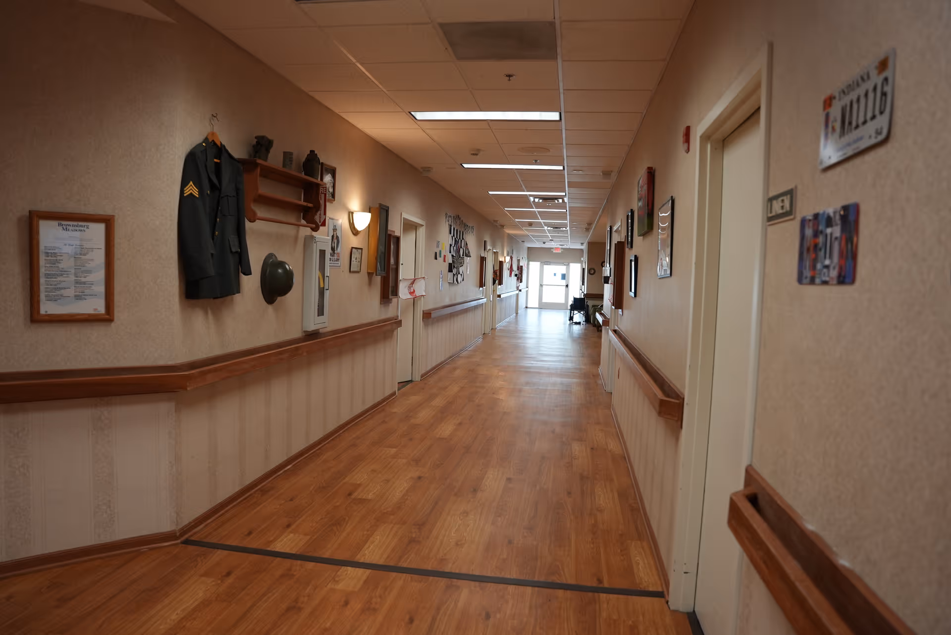 A long, well-lit hallway in Brownsburg Meadows facility with wooden flooring and beige walls. The walls are decorated with framed pictures, a military jacket, and a helmet. Handrails run along both sides of the hallway, and several doors are visible, including one labeled 'Men'. The hallway leads to a bright exit door at the far end.