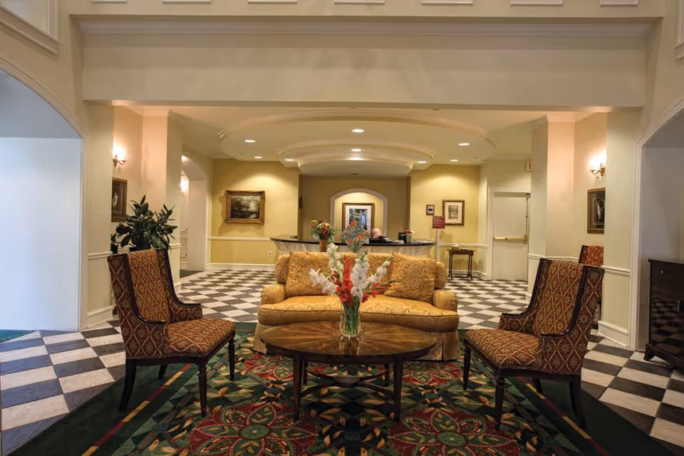 A well-lit interior lounge area with a yellow patterned sofa and two matching armchairs arranged around a wooden coffee table with a vase of flowers. The floor has a black and white checkered tile pattern with a decorative rug under the seating. The background shows a reception desk with a person seated behind it, framed artwork on the walls, and soft yellow walls with white trim.