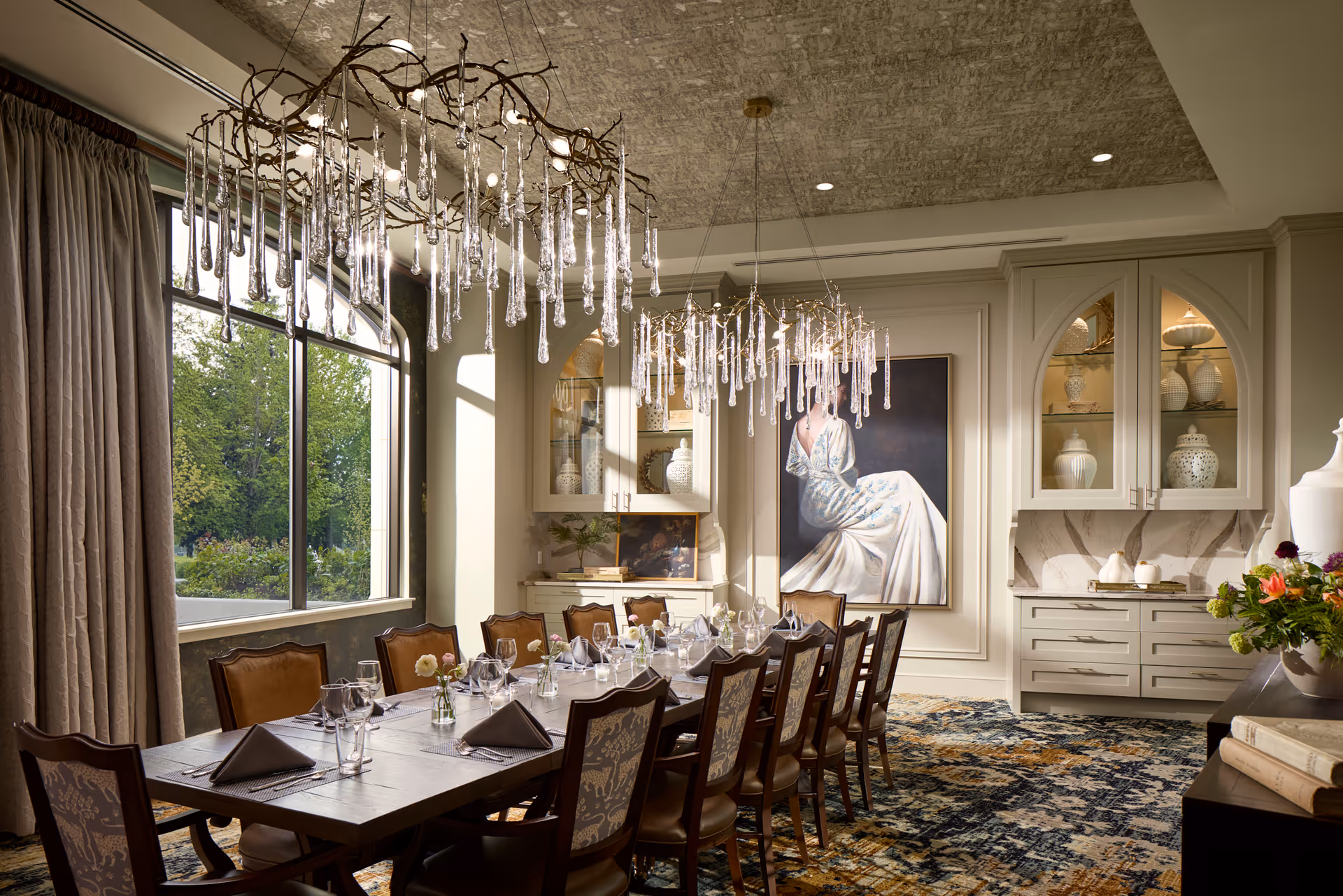 A formal dining room with a long wooden table set for a meal, surrounded by upholstered chairs. Two artistic chandeliers with hanging glass elements illuminate the room. Large windows with curtains allow natural light to enter, and decorative cabinets with vases and a large painting of a woman in a white dress adorn the walls. The room features a patterned carpet and elegant decor.