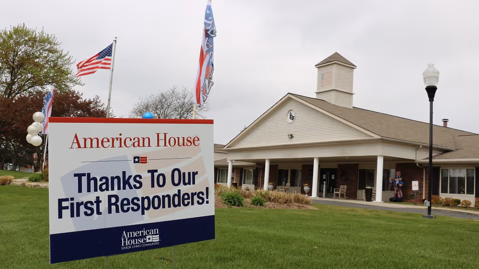 Exterior view of American House Sterling Heights senior living facility with a sign in the foreground that reads 'Thanks To Our First Responders!' The building has a covered entrance with white columns and a small tower on the roof. There are flags and balloons near the sign, and a lamp post on the right side of the image.