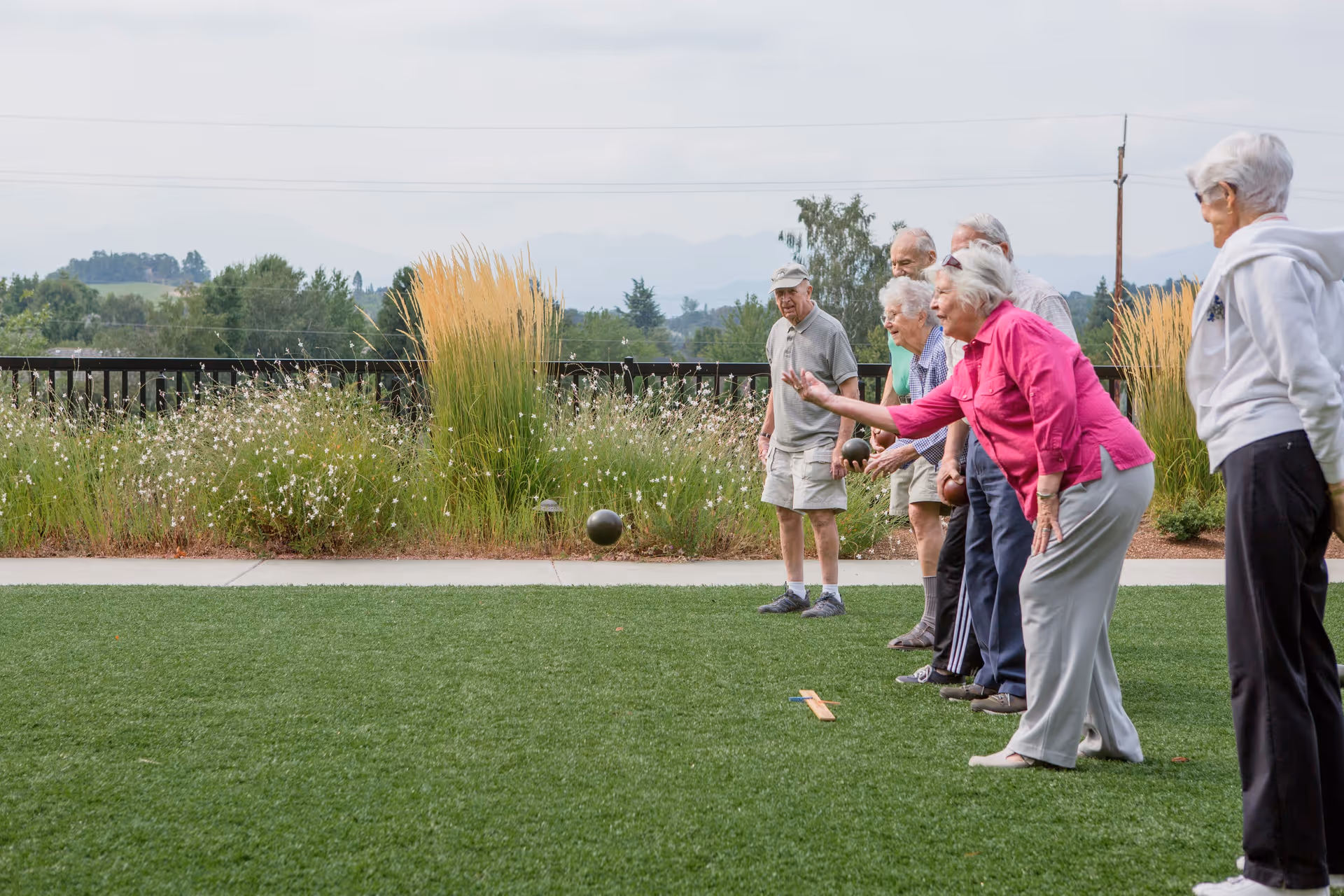 A group of elderly people playing bocce ball on a green lawn outdoors with plants and trees in the background under a cloudy sky.