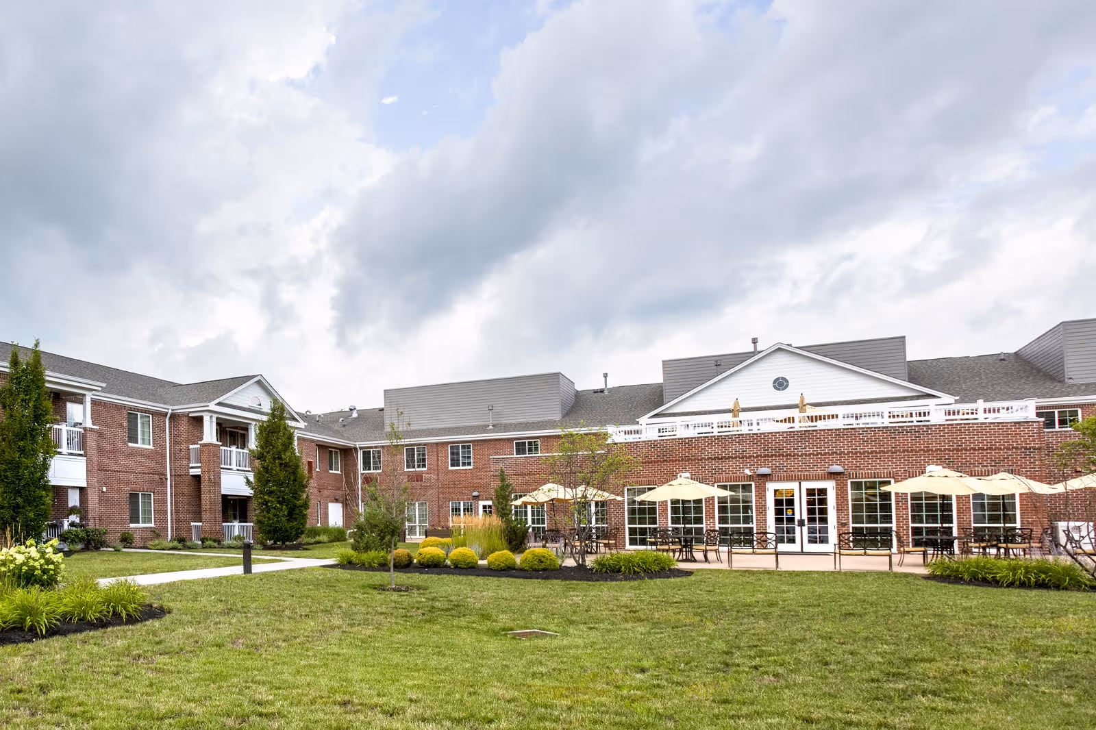 Front exterior of a two-story brick senior living building with a patio, umbrellas, and a landscaped lawn under a cloudy sky.