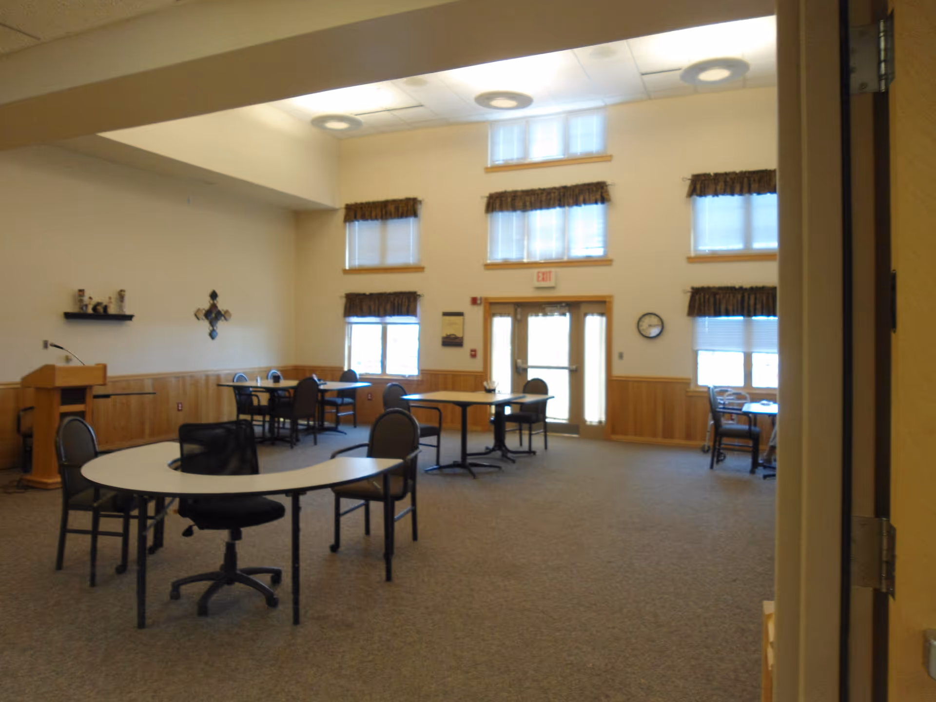 Spacious senior living common room with several tables and chairs, high windows, and a double-door exit.
