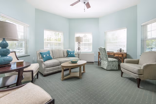 A bright and airy living room with light blue walls and multiple windows letting in natural light. The room features a beige sofa with blue accent pillows, two beige armchairs, a wooden coffee table, a side table with a blue lamp, a floor lamp, and a wooden desk with a patterned chair. The floor is carpeted in a muted green color with a subtle pattern, and a ceiling fan is visible above.