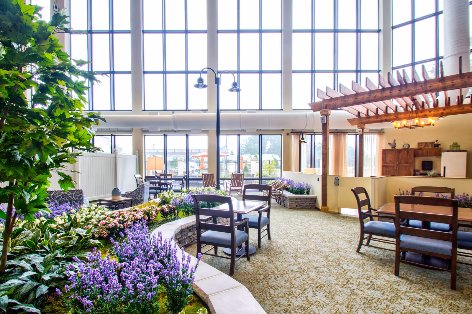 Bright and spacious indoor common area with large floor-to-ceiling windows letting in natural light. The room features several wooden tables and chairs, a variety of colorful flowers and plants arranged in stone-bordered garden beds, a wooden pergola structure with hanging lights, and a cozy seating area with armchairs. Outside the windows, patio umbrellas and outdoor seating are visible.