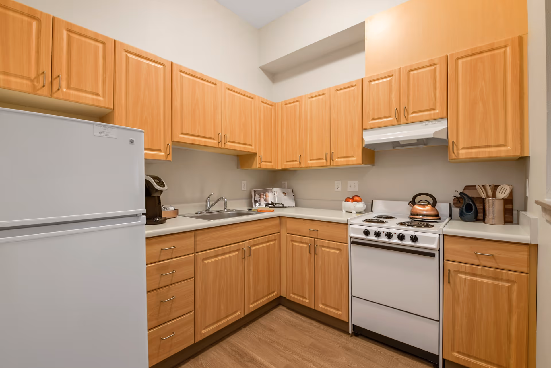 A compact kitchen with light wood cabinets, a white refrigerator, a white stove with a copper kettle on top, a stainless steel sink, a coffee maker, and various kitchen utensils on the countertop.