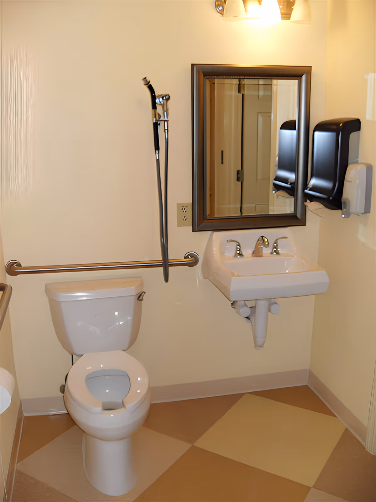 A clean and accessible bathroom with a white toilet, a wall-mounted sink with a mirror above it, two black soap or paper towel dispensers on the wall, a grab bar next to the toilet, and a handheld shower spray attached to the wall.