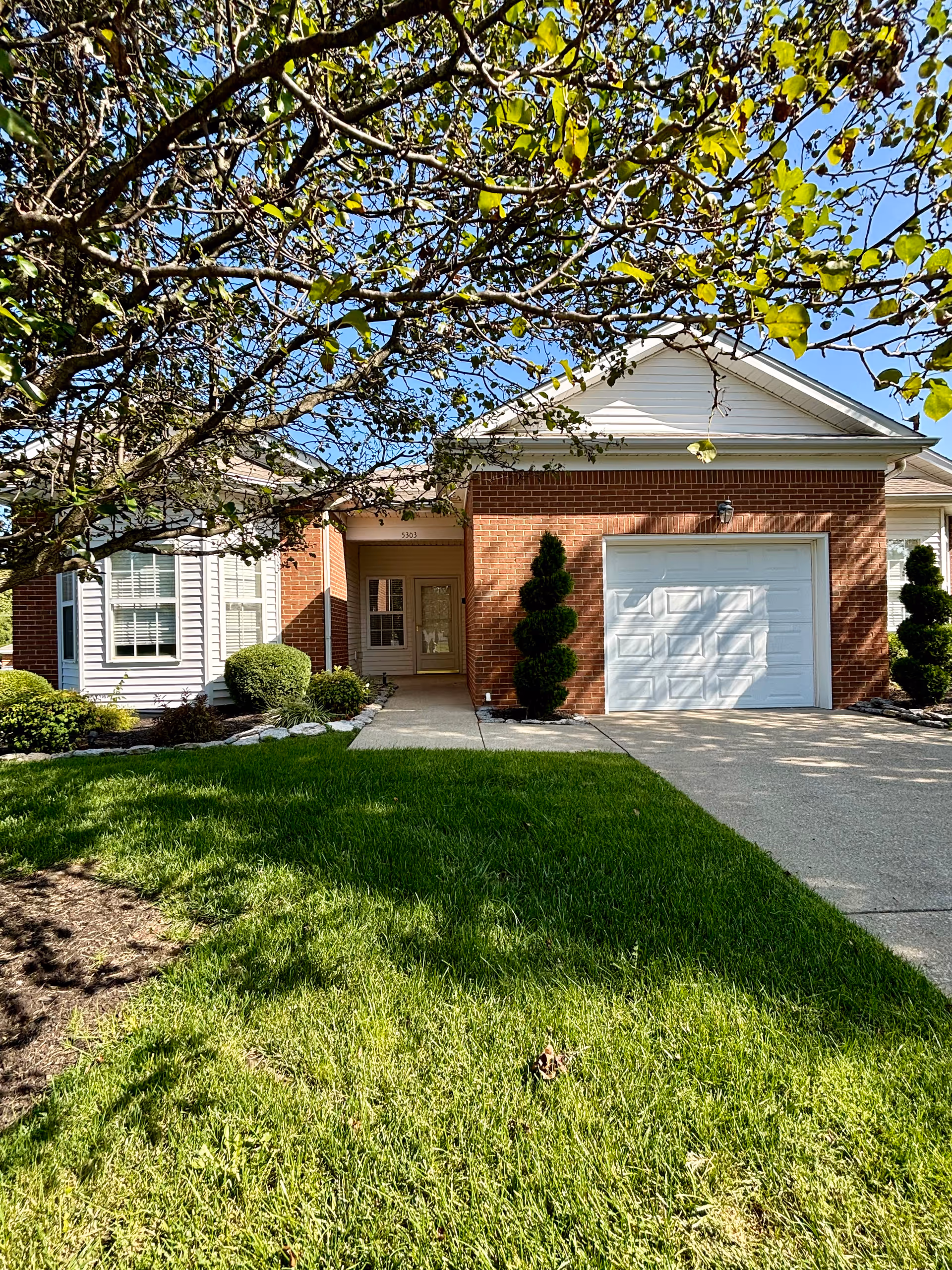 Front exterior view of a single-story brick and siding house with a white garage door, a concrete driveway, a walkway leading to the front door, neatly trimmed bushes, and a tree with green leaves partially shading the lawn.