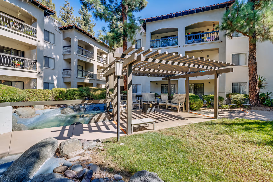 Outdoor courtyard area of a senior living facility with a pergola-covered seating area featuring wooden chairs and a fire pit. Surrounding the courtyard are multi-story residential buildings with balconies. There is a small water feature with rocks and greenery around the space, and tall trees provide shade.